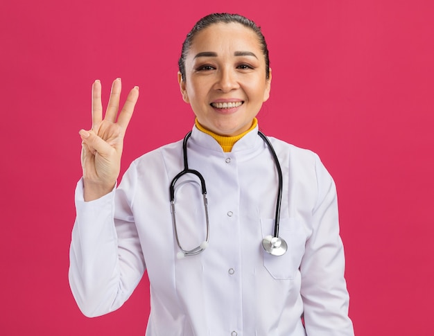 Молодая женщина-врач счастлива и позитивна, показывая номер три пальцами