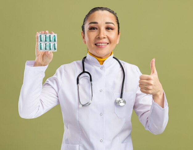 Young woman doctor   holding blister with pills   smiling confident showing thumbs up