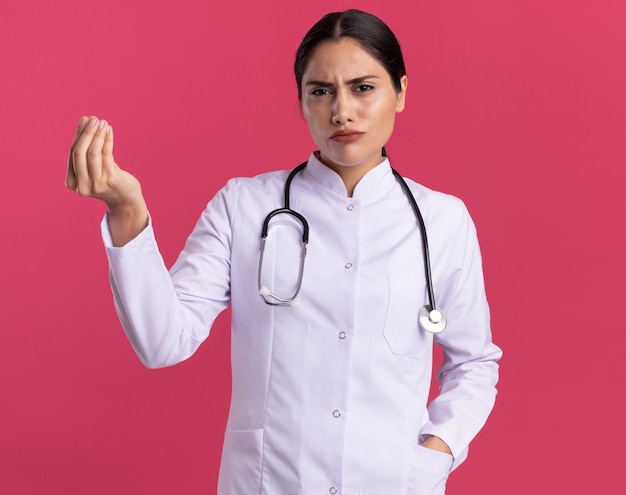 분홍색 벽 위에 서있는 손가락을 문지르는 돈 제스처를 보여주는 회의적인 표정으로 청진기와 의료 코트에 젊은 여자 의사