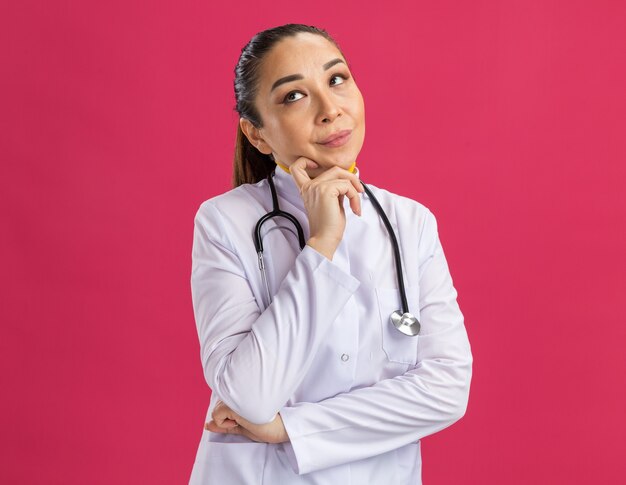 Young woman doctor   looking up with pensive expression with hand on her chin thinking