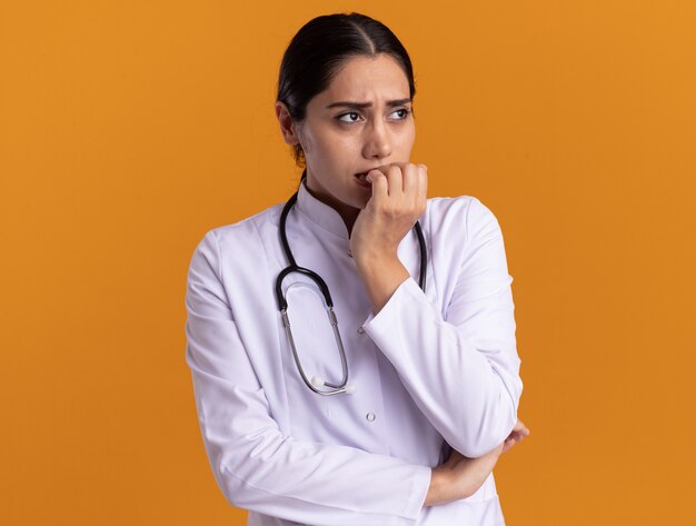Молодая женщина-врач в медицинском халате со стетоскопом на шее смотрит в сторону, напряженные и нервные кусающие ногти, стоящие над оранжевой стеной