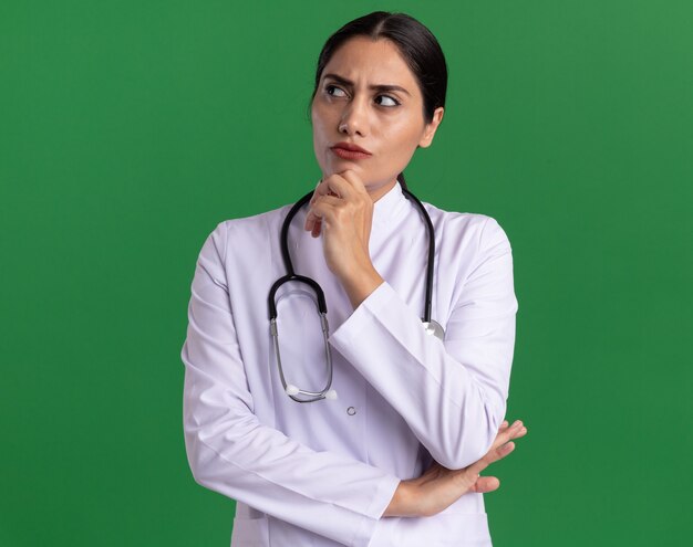 Young woman doctor in medical coat with stethoscope around her neck looking aside with pensive expression with hand on chin thinking standing over green wall