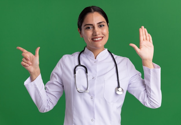 聴診器を首に巻いた医療コートを着た若い女性医師が、緑の壁の上に立って微笑んで人差し指で腕を上げる前を見て