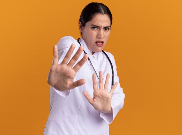 Молодая женщина-врач в медицинском халате со стетоскопом на шее, глядя вперед, испугалась, делая защитный жест руками, стоящими над оранжевой стеной