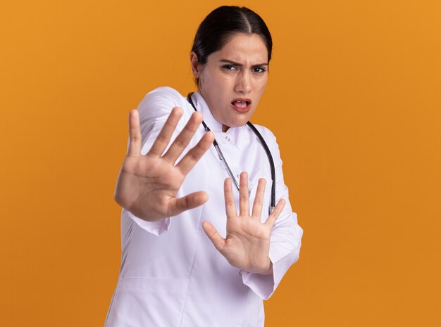 Young woman doctor in medical coat with stethoscope around her neck looking at front scared making defense gesture with hands standing over orange wall