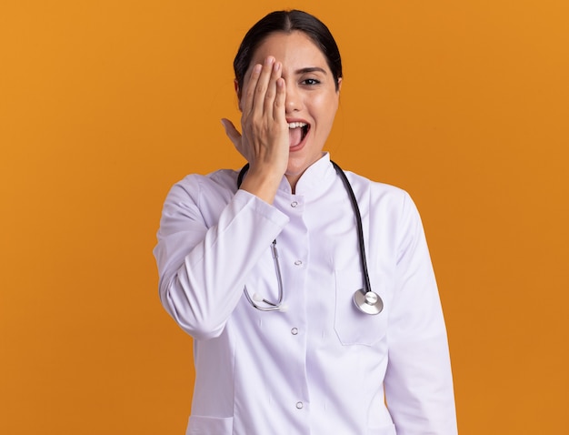 Young woman doctor in medical coat with stethoscope around her neck looking at front smiling with happy face covering eye with hand standing over orange wall