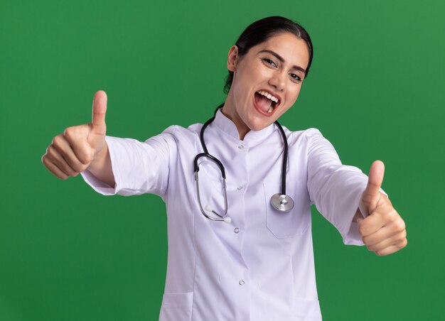 녹색 벽 위에 서있는 엄지 손가락을 유쾌하게 보여주는 행복한 얼굴로 정면을보고 그녀의 목 주위에 청진기와 의료 코트에 젊은 여자 의사