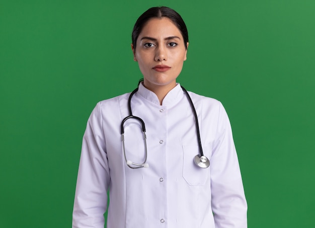 Young woman doctor in medical coat with stethoscope around her neck looking at front with serious confident expression standing over green wall