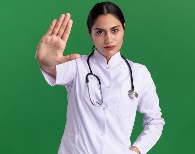 Молодая женщина-врач в медицинском халате со стетоскопом на шее, глядя вперед с серьезным лицом, показывая жест остановки с открытой рукой, стоящей над зеленой стеной