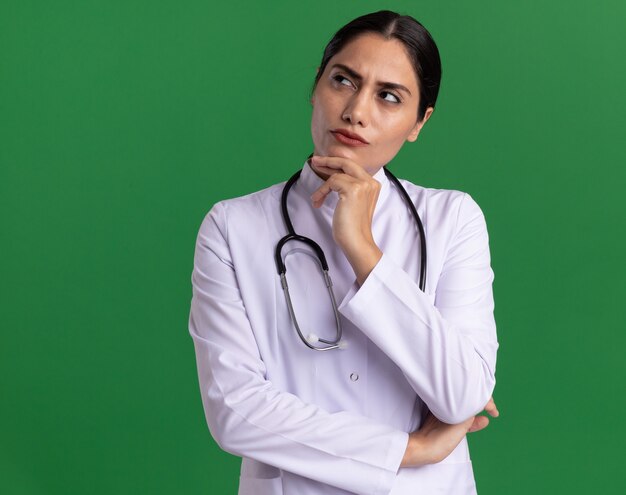 Молодая женщина-врач в медицинском халате со стетоскопом на шее, глядя вверх с задумчивым выражением лица, думая, стоя над зеленой стеной