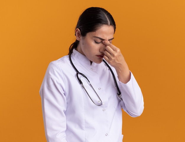 Молодая женщина-врач в медицинском халате со стетоскопом на шее, касающаяся носа между закрытыми глазами, усталая и скучающая, стоя над оранжевой стеной