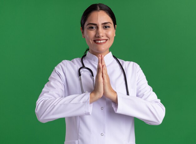 Young woman doctor in medical coat with stethoscope holding palms together like namaste gesture looking at front with smile on face standing over green wall