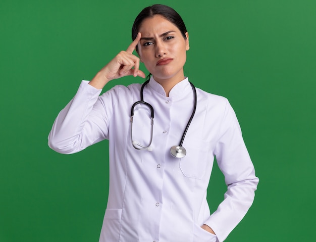 Young woman doctor in medical coat with stethoscope looking confused pointing with index finger at her temple standing over green wall