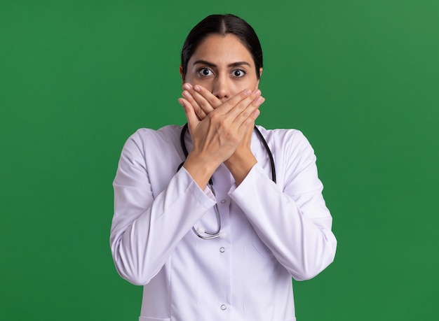 Young woman doctor in medical coat with stethoscope looking at front being shocked covering mouth with hands standing over green wall