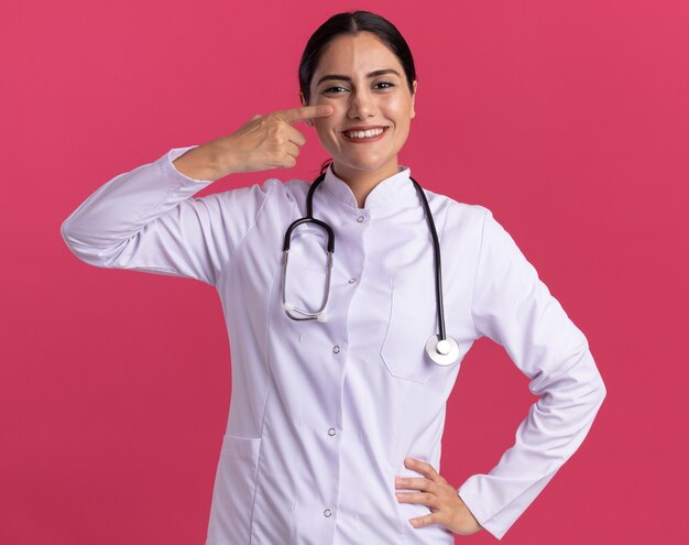 청진 기 의료 코트에 젊은 여자 의사 핑크 벽 위에 서 행복 한 얼굴로 웃 고 그녀의 눈에 검지 손가락으로 가리키는 앞