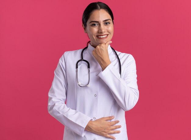 ピンクの壁の上に立って自信を持って笑顔の正面を見て聴診器と医療コートの若い女性医師