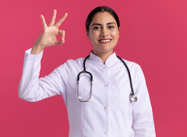 Young woman doctor in medical coat with stethoscope looking at front smiling with happy face showing ok sign standing over pink wall