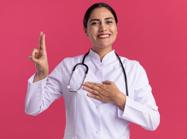 Free photo young woman doctor in medical coat with stethoscope looking at front swearing with hand on chest and fingers, making a loyalty promise smiling over pink wall