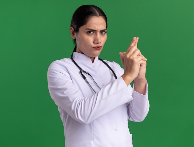 Foto gratuita medico della giovane donna in cappotto medico con lo stetoscopio che esamina anteriore con la faccia seria che fa il gesto della pistola con le dita che stanno sopra la parete verde