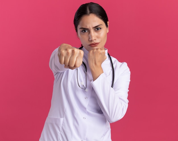 Молодая женщина-врач в медицинском халате со стетоскопом смотрит спереди с серьезным лицом, показывая сжатые кулаки спереди, стоя над розовой стеной