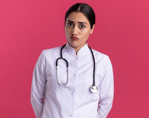 Young woman doctor in medical coat with stethoscope looking at front with serious face standing over pink wall