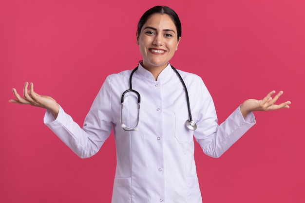 Young woman doctor in medical coat with stethoscope looking at front with smile on face with arms raised standing over pink wall