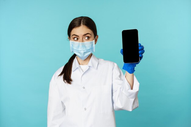 Young woman doctor in medical face mask and hospital uniform showing mobile phone app screen interfa...