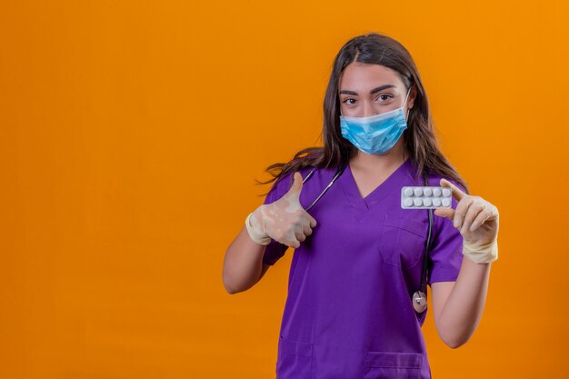 Young woman doctor in medical uniform with phonendoscope wearing protective mask and gloves smiling holding blister with pills showing thumb up over isolated orange background