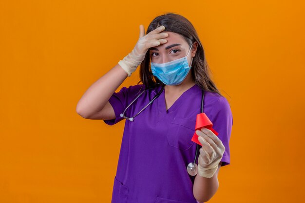 Young woman doctor in medical uniform with phonendoscope wearing protective mask and gloves smiling holding red ribbon and touching forehead with hand standing over isolated orange background