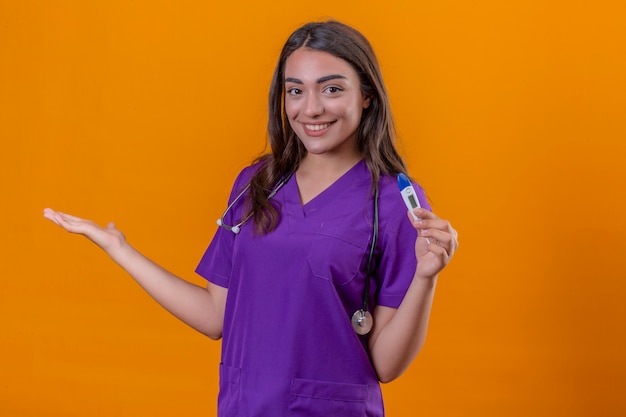 Young woman doctor in medical uniform with stethoscope smiling holding thermometer and with arm out in a welcoming gesture over isolated orange background