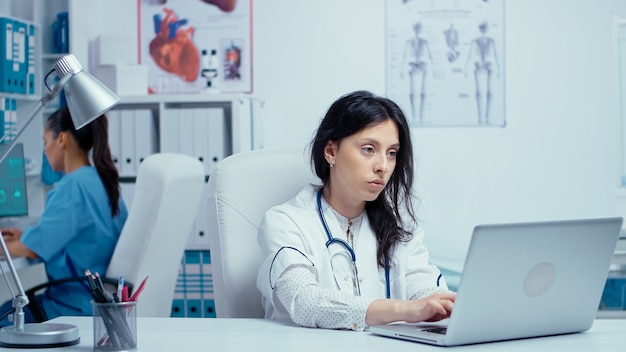 Young woman doctor in private medical cabinet typing on laptop while the nurse in working in the background. Healthcare system medical worker practitioner in hospital and health research