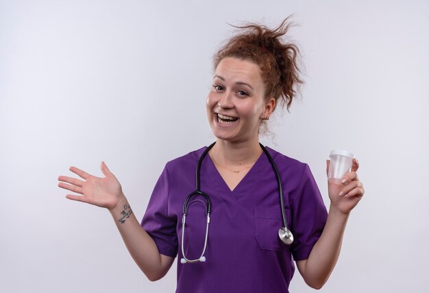 Young woman doctor wearing medical uniform with stethoscope holding test jar smiling with happy face raising arm standing over white wall