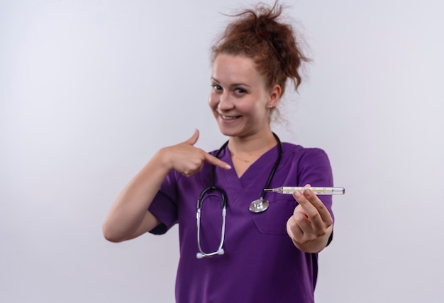白い壁の上に立って笑顔で自分自身に指で指している温度計を保持している聴診器で医療制服を着ている若い女性医師