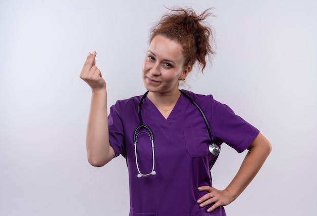 Foto gratuita medico della giovane donna che indossa l'uniforme medica con lo stetoscopio che sfrega le dita che fanno il gesto dei soldi con il sorriso sul fronte che sta sopra il muro bianco