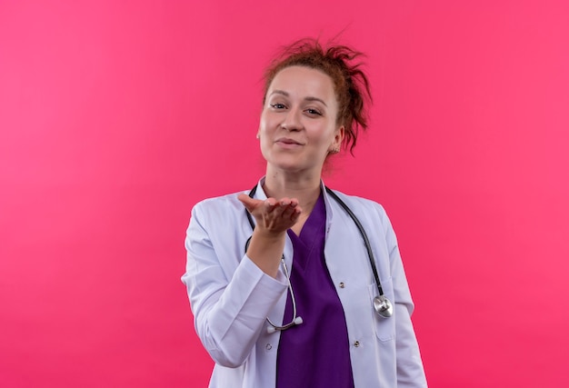 Giovane donna medico indossa camice bianco con lo stetoscopio che soffia un bacio con la mano in aria essendo incantevole in piedi sopra il muro rosa