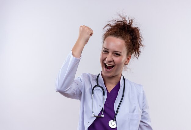 Young woman doctor wearing white coat with stethoscope clenching fist crazy happy rejoicing her success standing over white wall