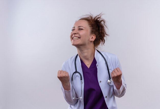 Young woman doctor wearing white coat with stethoscope clenching fists rejoicing her success happy and exited standing over white wall