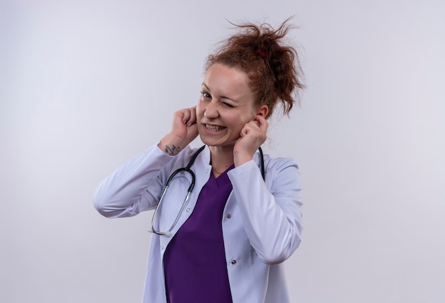 Young woman doctor wearing white coat with stethoscope closing ears with fingers with annoyed expression for the noise standing over white wall