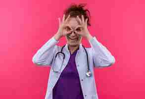 Foto gratuita giovane donna medico indossa camice bianco con uno stetoscopio facendo segno ok con le dita come un binocolo guardando attraverso le dita sporgenti lingua in piedi sopra la parete rosa