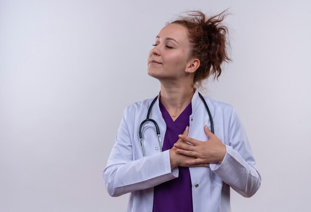 Giovane donna medico indossa camice bianco con lo stetoscopio che tiene le braccia sul petto sensazione di emozioni positive con gli occhi chiusi in piedi sul muro bianco