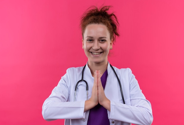 Medico della giovane donna che indossa camice bianco con lo stetoscopio che tiene le braccia insieme nel gesto di namaste sentirsi emozioni grate e positive sorridente in piedi sopra il muro rosa