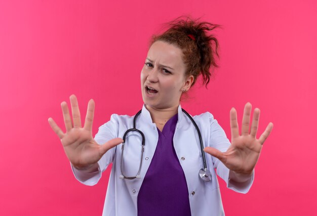 Young woman doctor wearing white coat with stethoscope holding open palms making stop sign with fear expression standing over pink wall