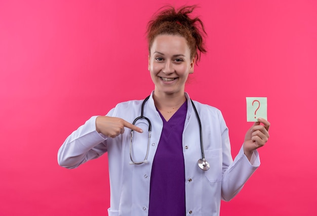 Giovane donna medico indossa camice bianco con uno stetoscopio tenendo la carta di promemoria con un punto interrogativo puntato con il dito sorridente allegramente in piedi sopra il muro rosa