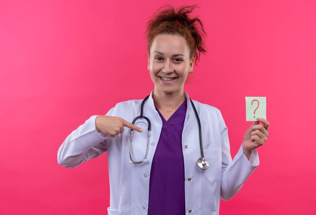 Young woman doctor wearing white coat with stethoscope holding reminder paper with question mark pointing with finger smiling cheerfully standing over pink wall