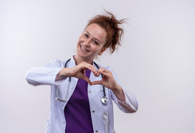 Молодая женщина-врач в белом халате со стетоскопом делает сердечный жест пальцами на груди, улыбаясь позитивно и счастливо, стоя над белой стеной