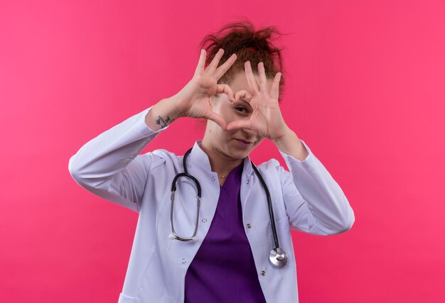 Молодая женщина-врач в белом халате со стетоскопом делает сердечный жест пальцами, глядя сквозь пальцы, улыбаясь, стоя над розовой стеной