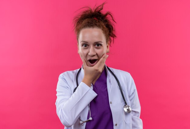 Young woman doctor wearing white coat with stethoscope shocked and surprised standing over pink wall