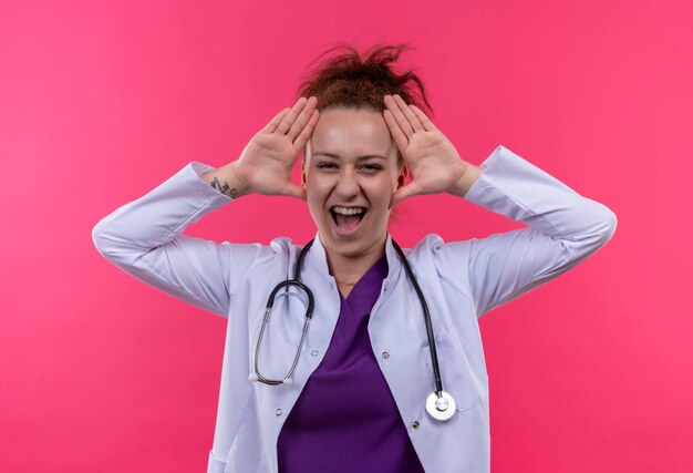 Молодая женщина-врач в белом халате со стетоскопом кричит с раздраженным выражением лица, держа руки возле головы, стоящей над розовой стеной