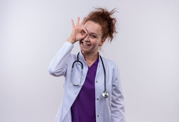 Young woman doctor wearing white coat with stethoscope smiling cheerfully doing ok sign looking through this sign standing over white wall