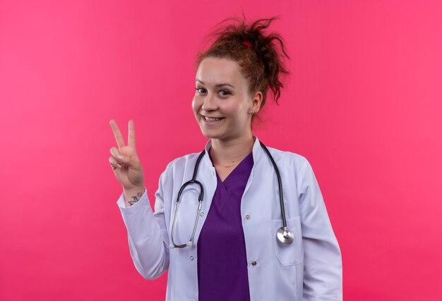 Молодая женщина-врач в белом халате со стетоскопом весело улыбается, показывая знак победы, стоящий над розовой стеной